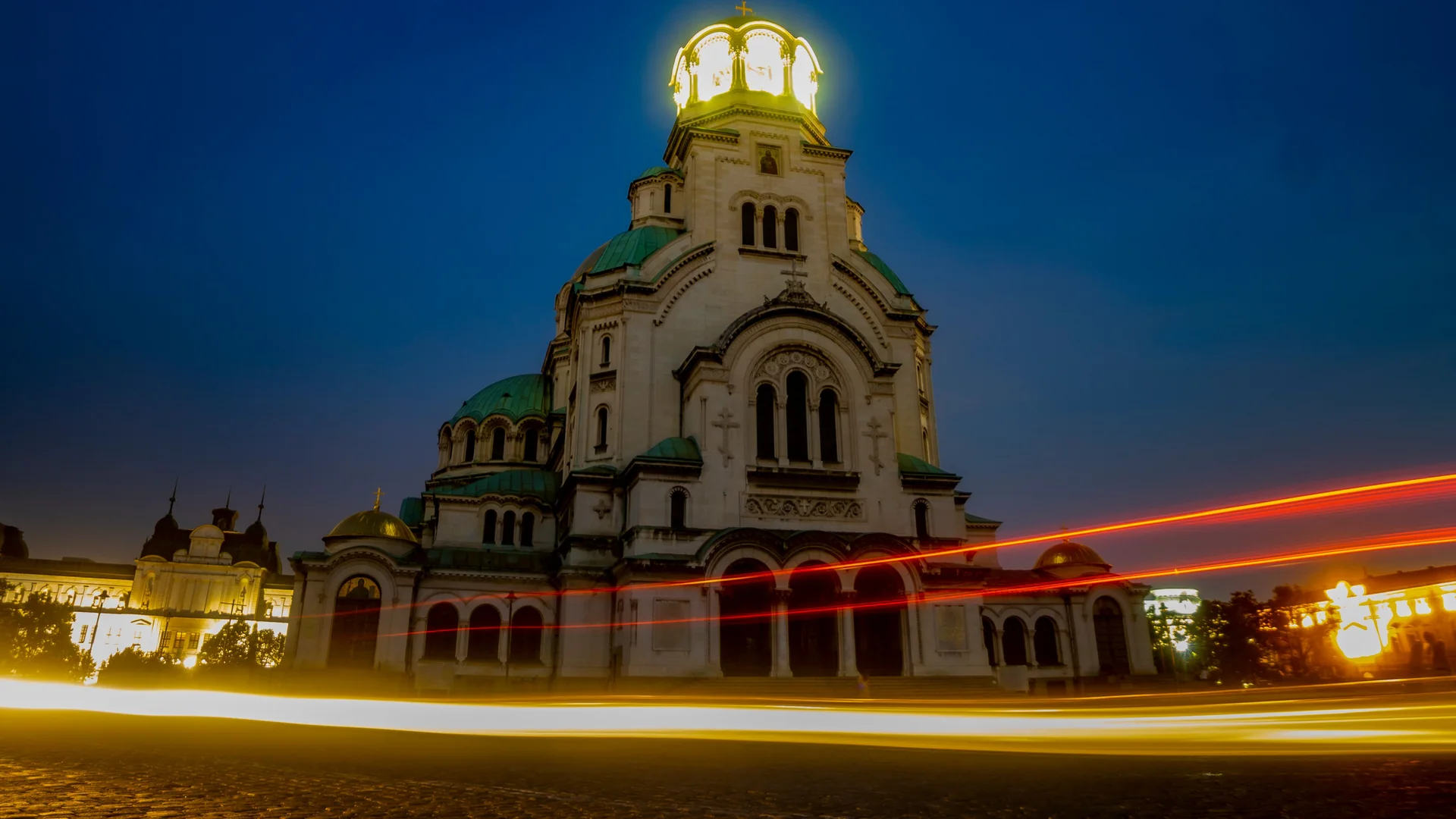 Alexander Nevsky Cathedral in Sofia lit gold at night, with light trails from traffic in the foreground